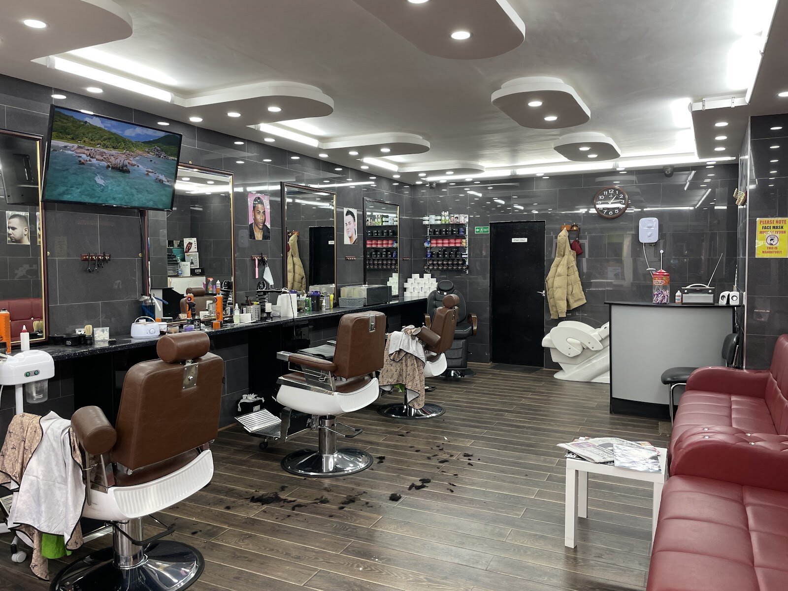 Inside the shop: four barber chairs down a wood floor with grey tiled walls, mirrors and a television on the wall, and a red leather bench along the far side to wait on.