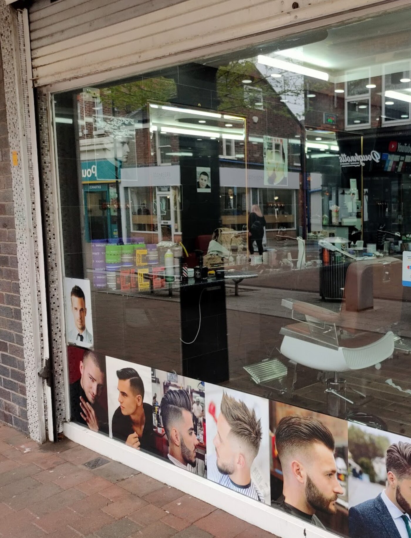 The shop window from the pavement, with a strip of haircut photographs along the bottom of the glass and the chairs and mirrors visible inside.