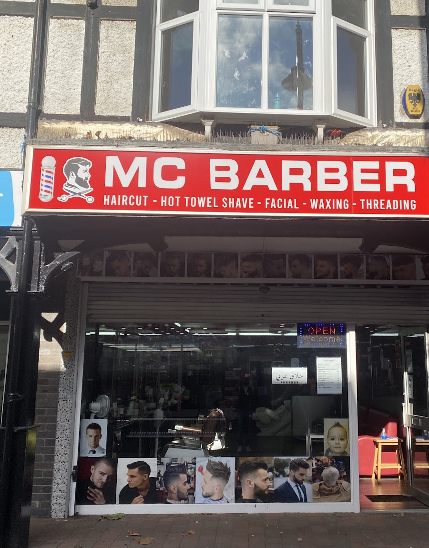 The front of the shop on High Road: a red fascia board reading MC BARBER in white capitals, with a barber pole and a bearded barber's head either side of the name, and the line “Haircut, hot towel shave, facial, waxing, threading” underneath it.